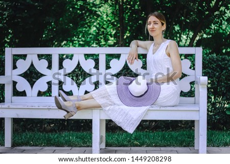 Young beautiful girl in a white dress sitting on a white bench in the summer park