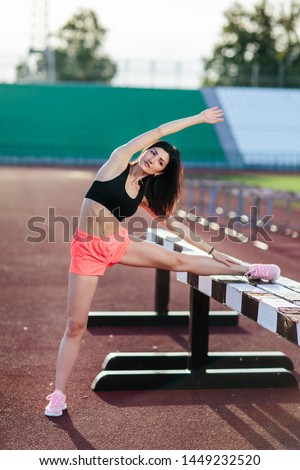 Beautiful brunette runner woman doing stretching leaning her leg on barrier for running stretching before workout - outdoor shot. Sport girl doing exercise in the stadium.