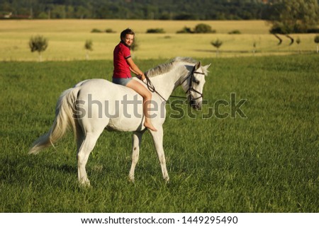Young girl ride on white horse without saddle on meadow in late afternoon, sunset time