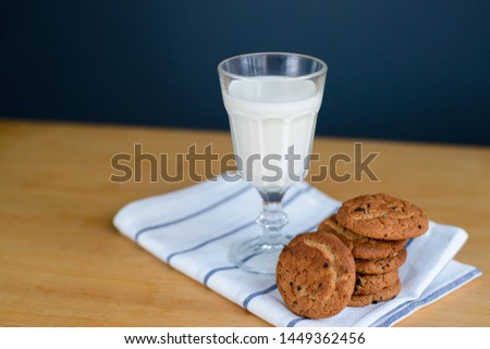round brown wholemeal oatmeal cookies stack with raisins and white milk in glass cup on striped textile rag on wooden table, close up side view of vertical still life stock photo image