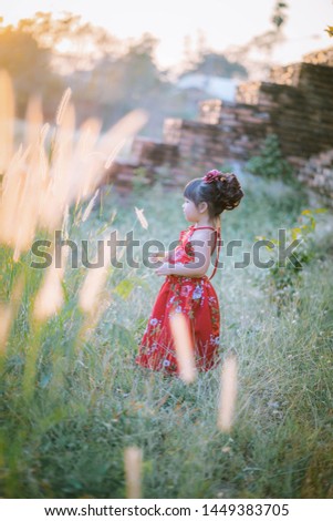 Asian little girl in red dress, walking like Happy in the flower garden and sunset. Outdoor Kids Fashion. Sweet toddler girl with floral head wreath on and flowers bunch in hands 