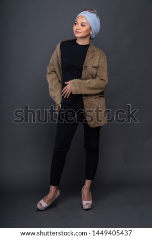 Gorgeous young woman dressed in black shirt with hijab, blazer and heeled shoes posing in studio. Beautiful Muslim girl demonstrating stylish smart clothing against grey wall on background.