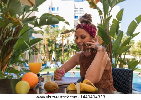 Colorful photo of a beautiful university girl taking notes while talking on the telephone on the terrace. Fruits and orange juice on the table. Banana trees and a blue swimming pool in the background