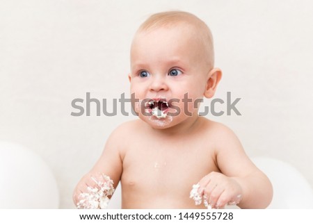 Baby birthday party. Infant eating birthday cake. Hungry boy on a light background with white ballons and copy space smash the cake.