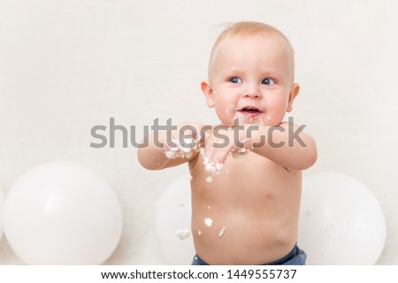Baby birthday party. Child eating birthday cake. The boy on a light background with white ballons and copy space smash the cake.