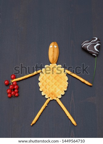 Funny little man with a chocolate heart ball and a branch of red berries made of cookies, waffles and breadsticks on a dark background. Delicious fun and entertainment for children in the kitchen.