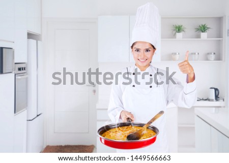 Picture of young female chef showing thumb up and tasty food in a pan in the kitchen