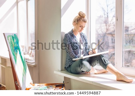 serious fai-haired girl with a pencil in her hair painting nature, landscape while resting on the windowsill. full length photo