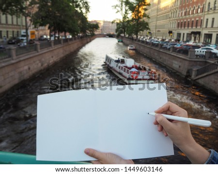 Mockup of person's hand holding blank white notebook preparing to write down his or hers ideas. Close up of female hand holding a pen and opened sketchbook pages on the city canal background 