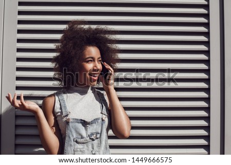 Young woman wearing denim overall shorts in the city street standing isolated on shutter answering phone call talking with boyfriend looking aside confused