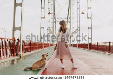 Full-lenght shot of stylish european woman wearing pink skirt and white jacket walking with a corgi dog in morning sunny city