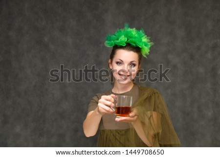 Portrait in lower chest on a gray background of a pretty young brunette woman with a green floral wreath in her hair. He stands with a cup of tea in his hands, talking, with emotions.