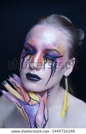 Perfect face young beautiful girl touching her face, bright make-up, purple orange yellow lines of paints flowing from face to hand, mascara tears flow from eyes. Long earrings, manicure black nails