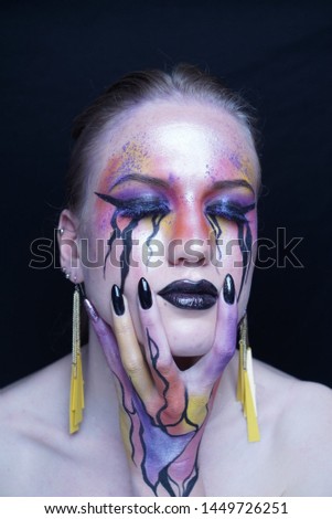 Perfect face young beautiful girl touching her face, bright make-up, purple orange yellow lines of paints flowing from face to hand, mascara tears flow from eyes. Long earrings, manicure black nails