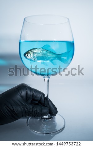 One hand in black glove holding a wine glass of fish in blue water
