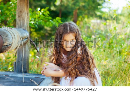 Little adorable girl with long hair in a gentle pink dress sitting near a well, summer day