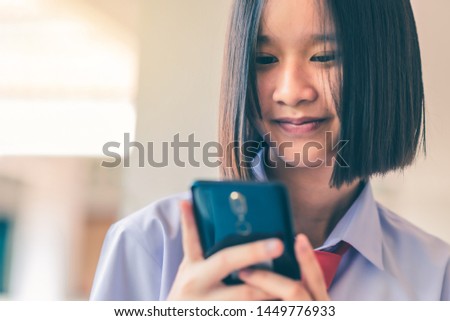 Smiling female Asian high school student in white uniform is enjoying social media on her smartphone.