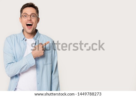 Surprised young man showing by forefinger at blank copy space. Stunned, amazed, astonished male standing pointing aside with index finger wearing glasses looking at camera isolated on gray background
