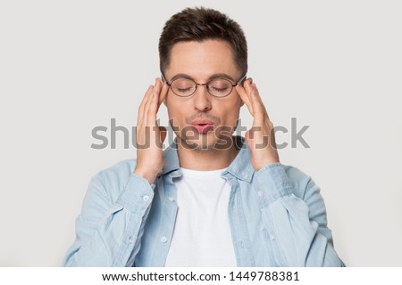 Young caucasian man in eyeglasses massaging temples with close eyes, breathing out, trying to calm down, posing on grey studio background head shot portrait, reducing stress, self-control concept. Royalty-Free Stock Photo #1449788381