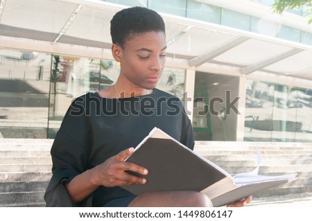 Serious focused black woman sitting on stairs outside and studying photo book. African American lady reading book or viewing folder with documents. Reader concept