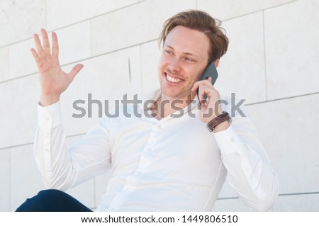 Smiling business man talking on phone and gesturing outdoors. Guy using cellphone and sitting with building wall in background. Communication in business concept.