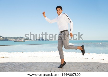 Cheerful young Latin man dancing on quay outdoors. Handsome man wearing shirt, looking camera, dancing or jumping on stone pavement with sea and blue sky in background. Happiness concept.
