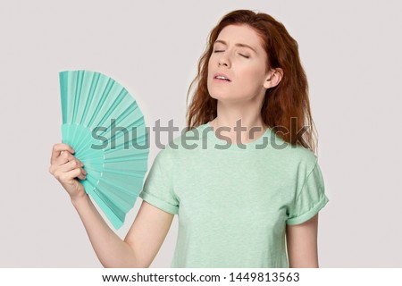 Overheated young red-haired woman using fan, cooling herself, suffering from hot summer weather or high temperature inside, heat stroke, feeling unwell, sweating, isolated on grey studio background. Royalty-Free Stock Photo #1449813563