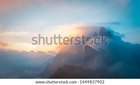 A stunning capture of the famous Seceda range covered in clouds and fog with a rising sun in the background. Blue and Orange cover the image of the alpine peaks in the Italian Dolomites.