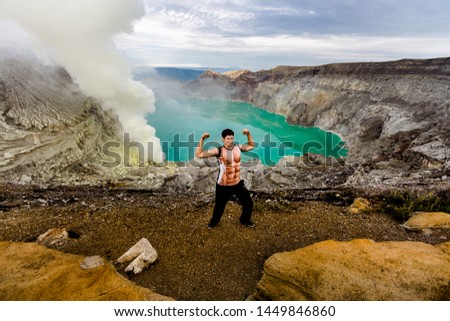 A young man is having fun around in the crater of a volcano against the background of a green sulfur lake and volcanic smoke.