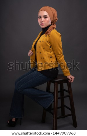 Fashionable female model in dark blue jeans, long sleeves yellow leather jacket with hijab isolated on grey background. Stylish Muslim hijab fashion lifestyle portraiture concept.
