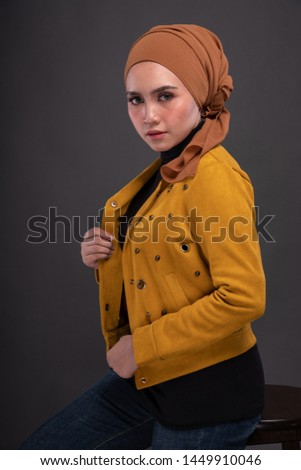 Fashionable female model in dark blue jeans, long sleeves yellow leather jacket with hijab isolated on grey background. Stylish Muslim hijab fashion lifestyle portraiture concept.