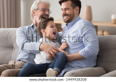 Happy grandfather and father tickling little boy, three generations of men having fun together, sitting on couch, laughing grandparent with son and grandchild relaxing spending weekend at home Royalty-Free Stock Photo #1449928208