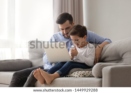 Caring father teaching adorable little son to use laptop, dad and preschool boy child looking computer screen, playing game, watching video or cartoons, sitting on couch together, family weekend