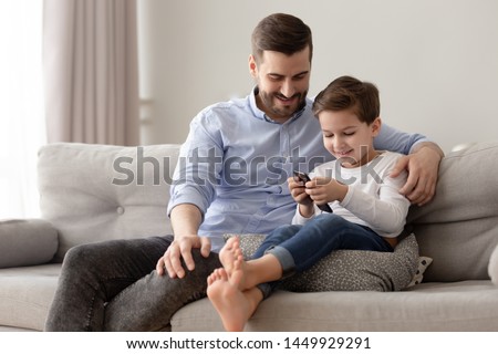 Smiling father and cute little son using phone apps together, watching cartoons or video, playing game, happy dad and preschool boy child sitting on couch, looking at mobile device cellphone screen