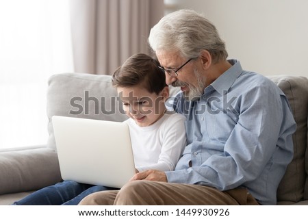 Happy grandfather and cute little grandson using laptop together, watching movie, playing game, sitting on couch at home, loving grandparent teaching preschool grandchild to use computer device