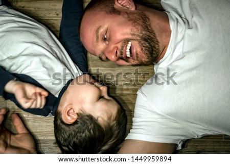 Young father and his beautiful son look at each other and smiling. Dad and son having fun at home. Handsome father and cute son laying on wooden floor and enjoying time together. Family bonding. 