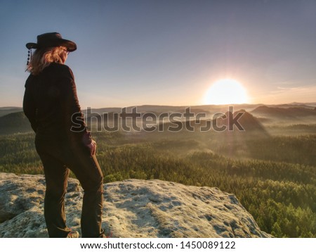 Beautiful long blond hair woman wearing cowgirl hat and dark trekking clothes  while resting in sunlit on summit.