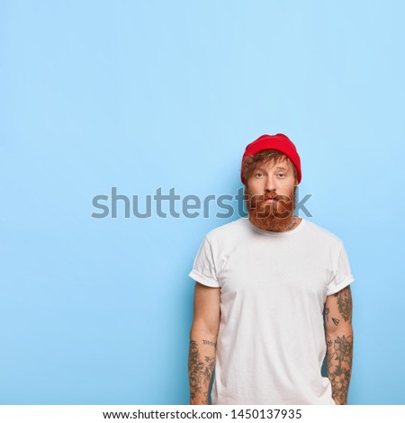 Serious good looking male model wears fashionable red hat and casual t shirt, going to have stroll, has tattooed arm, ginger beard, poses against blue wall with blank copy space on left side