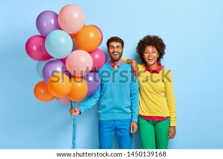 Young couple come on party, man holds bunch of many colorful balloons, his girlfriend stands near, wear bright casual clothes, isoated on blue background. Diverse boyfriend and girlfriend celebrate