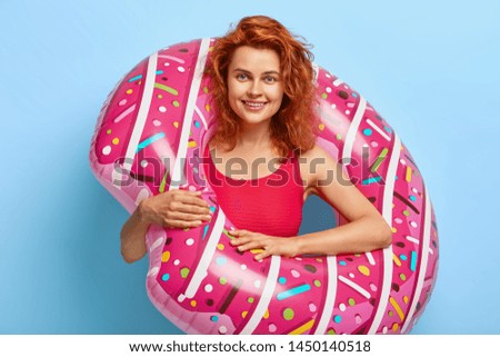 Beautiful relaxed red haired woman wears red bikini, stands inside of inflated swimring, looks with smile at camera, has awesome summer vacation in paradise place, isolated on blue background