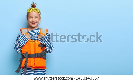 Happy little female kid wears goggles and lifevest, plays on beach, has good vacation, dressed in striped sweater, stands against blue background, blank space for your information. Summer holiday