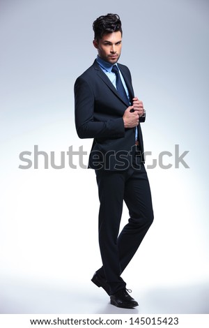 full length picture of a young business man posing with his hands on his suit jacket and looking at the camera. on a gray background