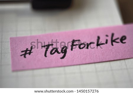 Popular hashtag "TagForLike" written on pink sticky note. It is stuck on the notepad with a pen and clipboard.