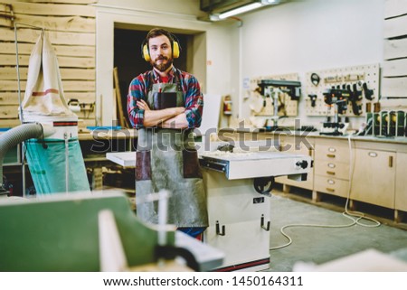 Half length portrait of self-employed woodman in work apron standing in own manufacturing fabric with crossed hands and looking at camera,bearded joiner in protective haedphones for noise cancellation Royalty-Free Stock Photo #1450164311