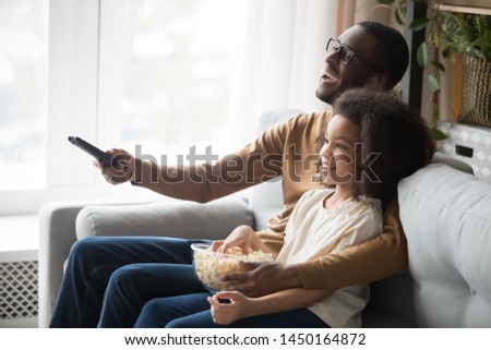 Smiling african American young dad relax on couch with cute preschooler daughter watching TV together, happy black father sit rest on sofa with girl child use remote enjoy movie time with popcorn