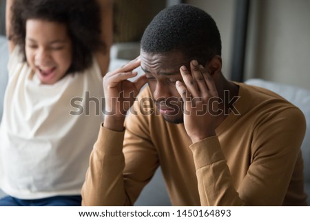Tired african american single dad touch massage temples suffer from severe headache or dizziness, happy disobedient small daughter have fun playing scream making noise at background. Fatigue concept Royalty-Free Stock Photo #1450164893