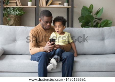 African American young dad and biracial preschooler child spend time together watch football on smartphone, surprised black father and little son sit on couch enjoy cartoon or video on cellphone