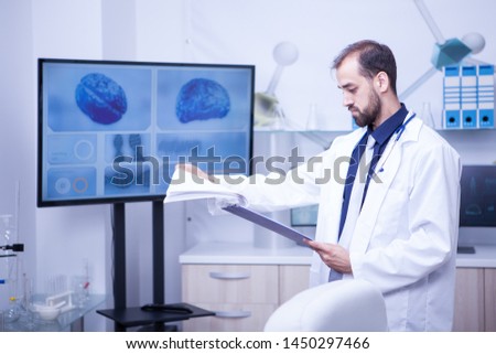 Young doctor turning pages with analyze on his clipboard in a laboratory for brain problems. Brain displayed on monitor.