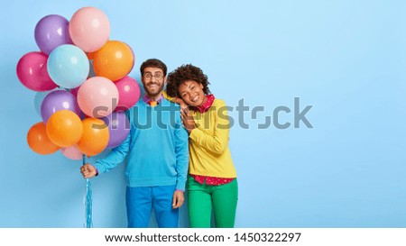 Horizontal shot of happy birthday man holds colorful balloons, Afro girl leans on his shoulder, wait for guests together, have broad smiles, isolated on blue wall with blank space. People and holiday