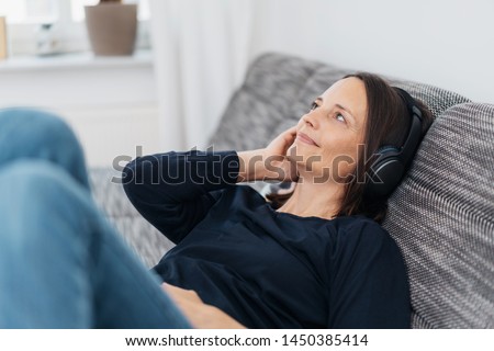 Happy adult woman listening to black headphones while leaning back on dark grey couch and wearing black shirt
